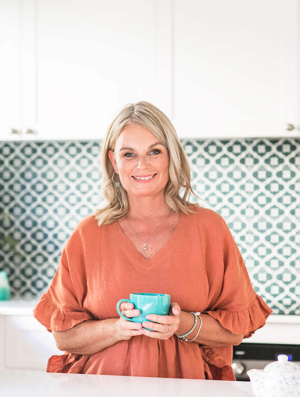 Rachel Scoltock Angel Medium holding a cup of tea and wearing a terracotta coloured top in an aqua kitchen